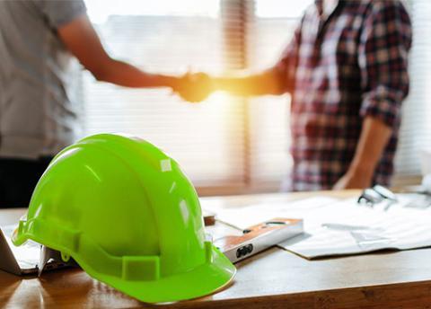 Two men shake hands with a desk in foreground with green hard hat.